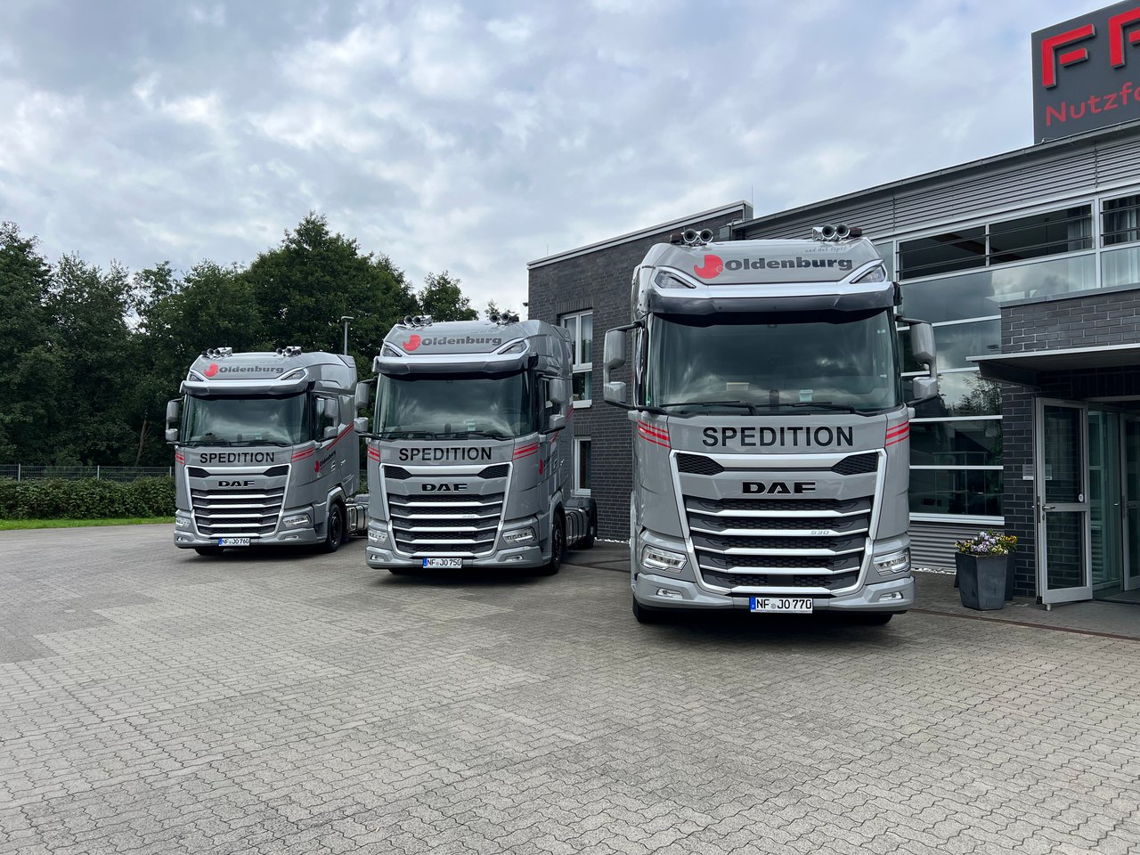 Drei graue DAF-Lkw mit "SPEDITION" und "Oldenburg" Aufschrift stehen vor einem Gebäude.